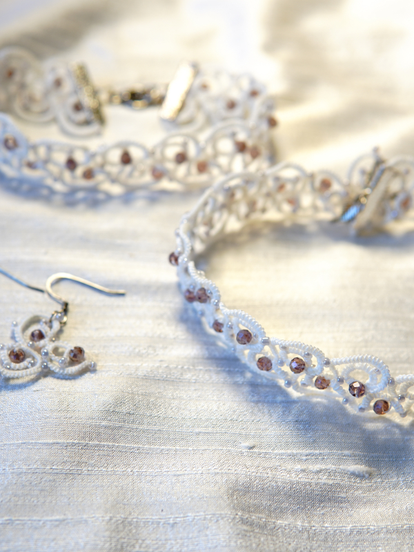 The Elarian Bloom Earrings shimmer with pale rose crystals in white tatted lace. Tatted earrings beside other jewelry in the set