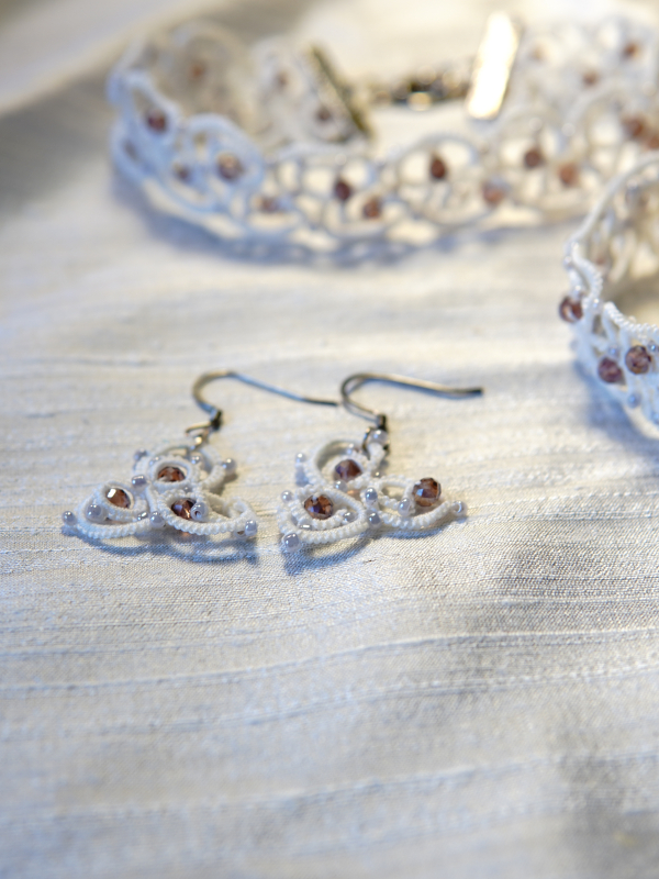 The Elarian Bloom Earrings shimmer with pale rose crystals in white tatted lace. Earrings laid beside matching necklace.