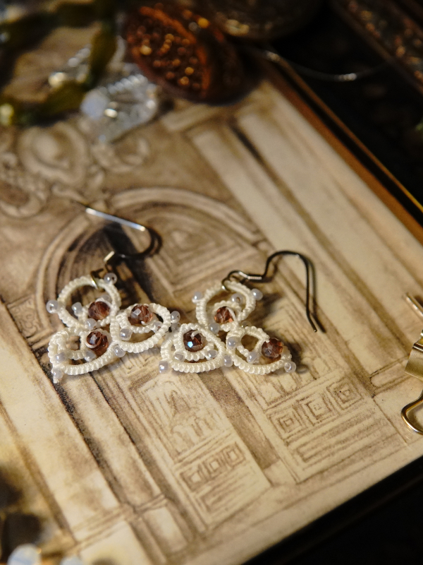 The Elarian Bloom Earrings shimmer with pale rose crystals in white tatted lace. White lace earrings resting on a framed backdrop