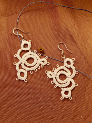 Close-up of handmade tatted lace earrings named Whirlwind against warm fabric.